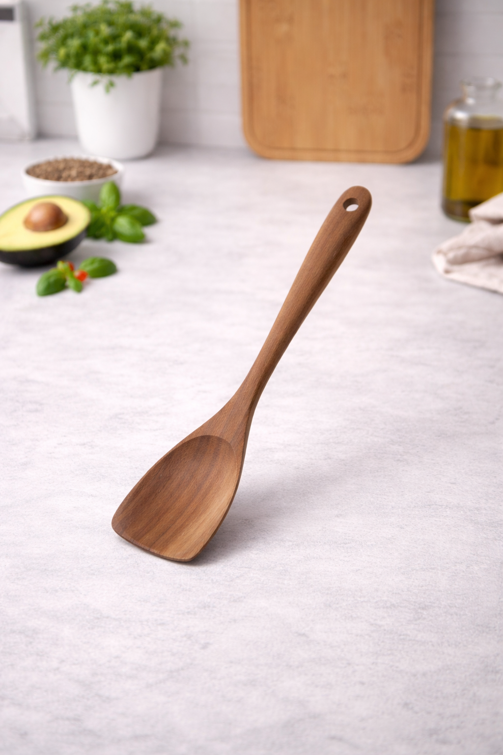 Wooden spoon on a kitchen counter with an avocado, leaves, and a cutting board in the background.